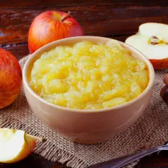Un bol de compote de pommes maison, entouré de pommes fraîches et de bâtons de cannelle, sur une table en bois rustique.