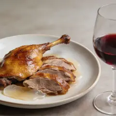 Un plat de confit de canard, coupé en tranches, servi avec une sauce légère, accompagné d'un verre de vin rouge.