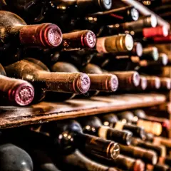 Des bouteilles de vin anciennes, poussiéreuses, sur des étagères en bois. Certaines ont des capsules rouges, d'autres dorées. On imagine le goût d'un vin qui pique en bouche.