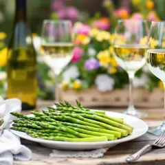 Un plateau d'asperges vertes fraîches, une bouteille de vin blanc et deux verres remplis de vin, sur une table en bois avec des fleurs en arrière-plan. La question quel vin avec asperges est parfaitement illustrée.