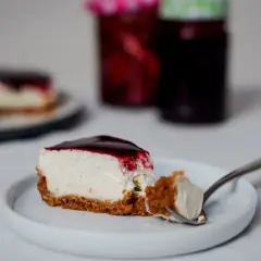 Une part de cheesecake sans cuisson crémeux, nappé de coulis de fruits rouges, sur une assiette blanche. Une bouchée est prélevée à la fourchette.