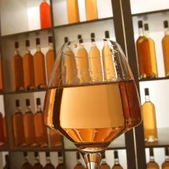 Verre de vin à la couleur rose très clair, devant une étagère remplie de bouteilles dorées.