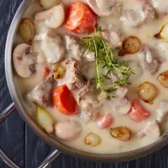 Un plat de blanquette de veau crémeuse avec des morceaux de viande tendre, des carottes, des champignons et des petits oignons, garni de thym frais.