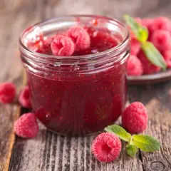 Un pot de confiture de framboise maison, garni de fruits frais et de feuilles de menthe, sur un fond de bois rustique.