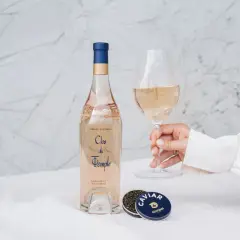 Une main tient un verre de vin rosé. À côté, une bouteille de Clos du Temple et une boîte de caviar. Une belle conservation rosé pour un moment d'exception.