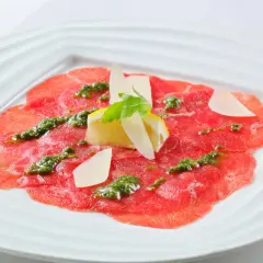 Carpaccio de bœuf finement tranché, arrosé d'une sauce verte et garni de copeaux de parmesan, de citron et d'une feuille de roquette.