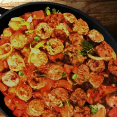 Un plat de rougail saucisse mijote dans une cocotte noire, avec des morceaux de saucisses, des tomates et des oignons.