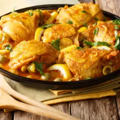 Un plat de poulet aux olives, citron et herbes fraîches, servi dans un plat noir avec des cuillères en bois.