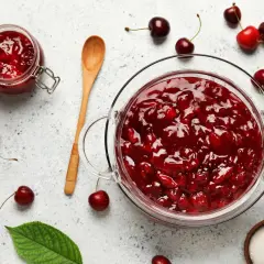 Un bocal et un bol en verre débordent de confiture de cerises maison. Des cerises fraîches et des feuilles vertes parsèment la surface.