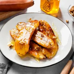 Bananes flambées caramélisées, nappées de miel et parsemées de noix, sur une assiette blanche. Un délice sucré.