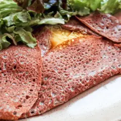 Une galette sarrasin pliée, garnie de fromage fondant et de salade verte fraîche, sur une assiette beige.