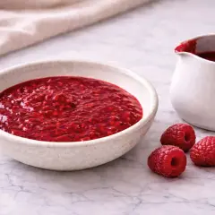 Un bol et un pichet remplis de coulis de framboise, avec trois framboises fraîches à côté.