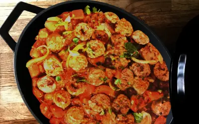 Un plat de rougail saucisse mijote dans une cocotte noire, avec des morceaux de saucisses, des tomates et des oignons.