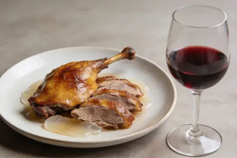 Un plat de confit de canard, coupé en tranches, servi avec une sauce légère, accompagné d'un verre de vin rouge.
