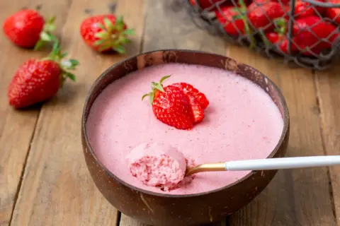Délicieuse mousse fraise dans une noix de coco, garnie d'une fraise fraîche. Une cuillère dorée en extrait une bouchée aérienne.