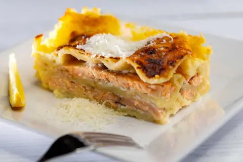 Une part généreuse de lasagne saumon, dorée au four, garnie de parmesan râpé et accompagnée d'un quartier de citron.