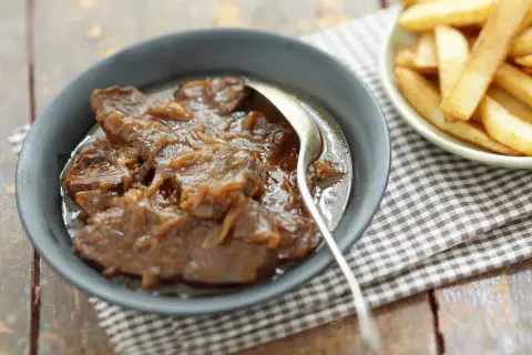 Un plat de carbonade flamande mijotée, servie dans un bol avec une cuillère, à côté d'une coupelle de frites dorées.
