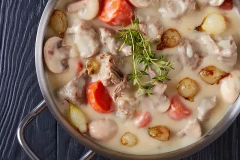 Un plat de blanquette de veau crémeuse avec des morceaux de viande tendre, des carottes, des champignons et des petits oignons, garni de thym frais.