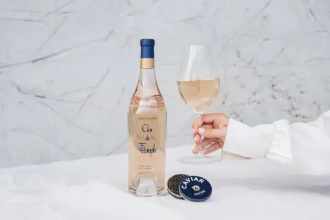 Une main tient un verre de vin rosé. À côté, une bouteille de Clos du Temple et une boîte de caviar. Une belle conservation rosé pour un moment d'exception.