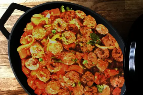 Un plat de rougail saucisse mijote dans une cocotte noire, avec des morceaux de saucisses, des tomates et des oignons.
