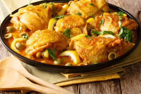 Un plat de poulet aux olives, citron et herbes fraîches, servi dans un plat noir avec des cuillères en bois.