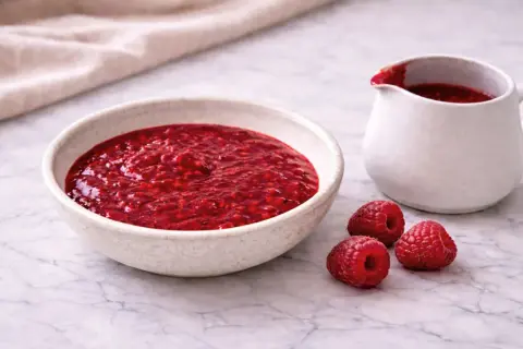 Un bol et un pichet remplis de coulis de framboise, avec trois framboises fraîches à côté.