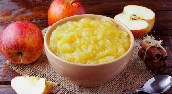 Un bol de compote de pommes maison, entouré de pommes fraîches et de bâtons de cannelle, sur une table en bois rustique.