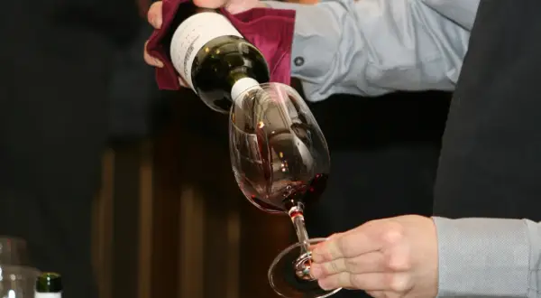 Un serveur verse du vin rouge dans un verre. Le **dépôt de vin blanc** sur la bouteille est visible.