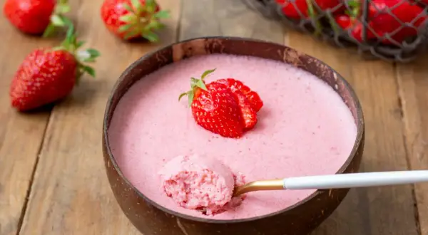 Délicieuse mousse fraise dans une noix de coco, garnie d'une fraise fraîche. Une cuillère dorée en extrait une bouchée aérienne.