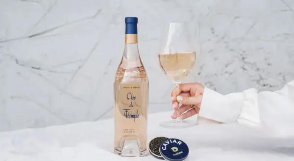 Une main tient un verre de vin rosé, à côté d'une bouteille et de caviar. Le danger du vin rosé est qu'il est si délicieux qu'on en redemande.
