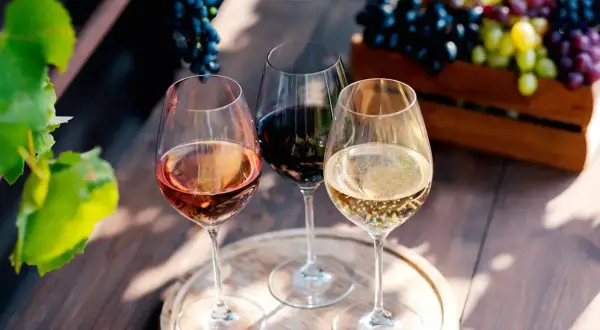 Trois verres de vin, un rosé, un rouge et un blanc pétillant, évoquent la dégustation de grands crus bordeaux, avec des grappes de raisin en arrière-plan.