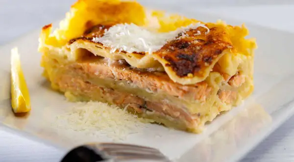 Une part généreuse de lasagne saumon, dorée au four, garnie de parmesan râpé et accompagnée d'un quartier de citron.