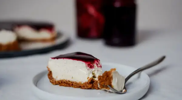 Une part de cheesecake sans cuisson crémeux, nappé de coulis de fruits rouges, sur une assiette blanche. Une bouchée est prélevée à la fourchette.