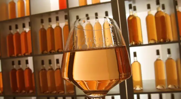 Verre de vin à la couleur rose très clair, devant une étagère remplie de bouteilles dorées.