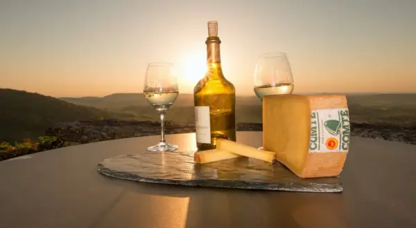 Un coucher de soleil sur le Jura, avec un verre de vin jaune, une bouteille et du fromage Comté. Qu'est-ce que le vin jaune ? C'est un vin du Jura, unique et savoureux.