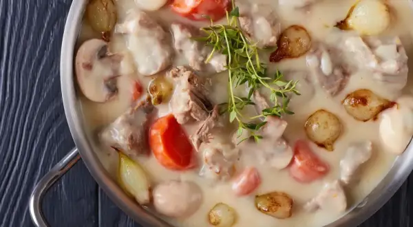 Un plat de blanquette de veau crémeuse avec des morceaux de viande tendre, des carottes, des champignons et des petits oignons, garni de thym frais.