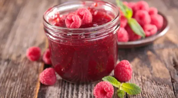 Un pot de confiture de framboise maison, garni de fruits frais et de feuilles de menthe, sur un fond de bois rustique.