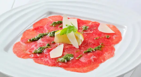 Carpaccio de bœuf finement tranché, arrosé d'une sauce verte et garni de copeaux de parmesan, de citron et d'une feuille de roquette.