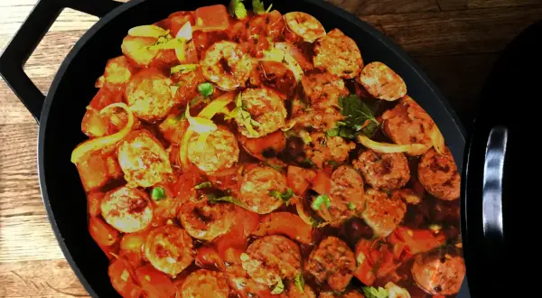 Un plat de rougail saucisse mijote dans une cocotte noire, avec des morceaux de saucisses, des tomates et des oignons.