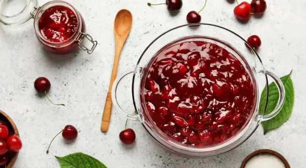 Un bocal et un bol en verre débordent de confiture de cerises maison. Des cerises fraîches et des feuilles vertes parsèment la surface.