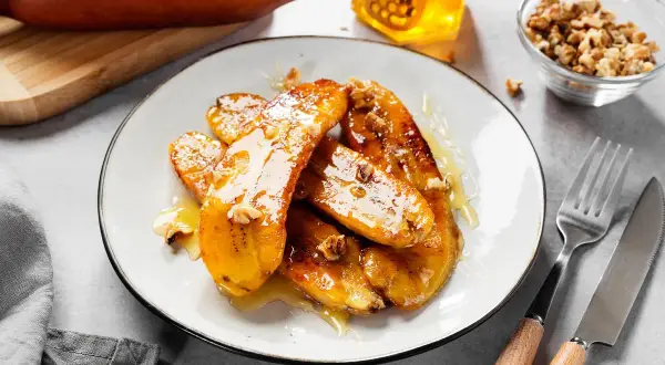 Bananes flambées caramélisées, nappées de miel et parsemées de noix, sur une assiette blanche. Un délice sucré.