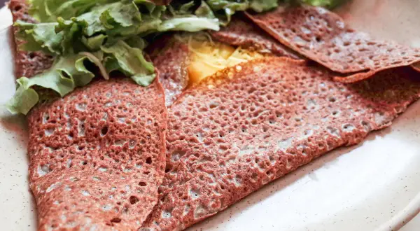 Une galette sarrasin pliée, garnie de fromage fondant et de salade verte fraîche, sur une assiette beige.
