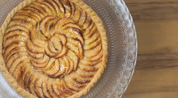 Une magnifique tarte aux pommes, aux pommes caramélisées disposées en spirale sur une pâte dorée, servie sur un plat en verre décoré.
