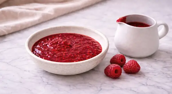 Un bol et un pichet remplis de coulis de framboise, avec trois framboises fraîches à côté.
