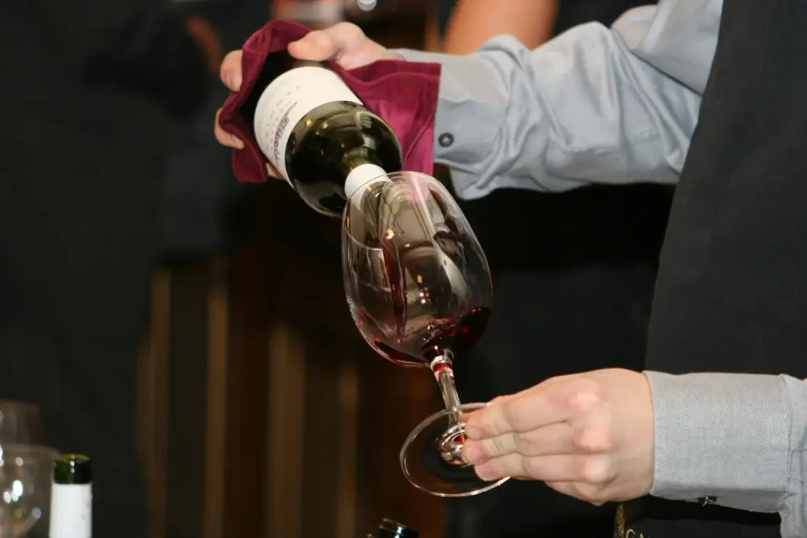Un serveur verse du vin rouge dans un verre. Le **dépôt de vin blanc** sur la bouteille est visible.