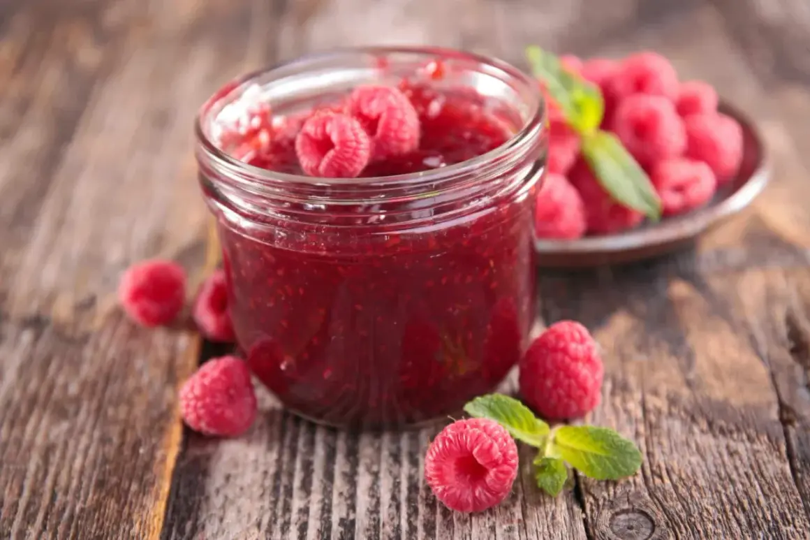 Un pot de confiture de framboise maison, garni de fruits frais et de feuilles de menthe, sur un fond de bois rustique.