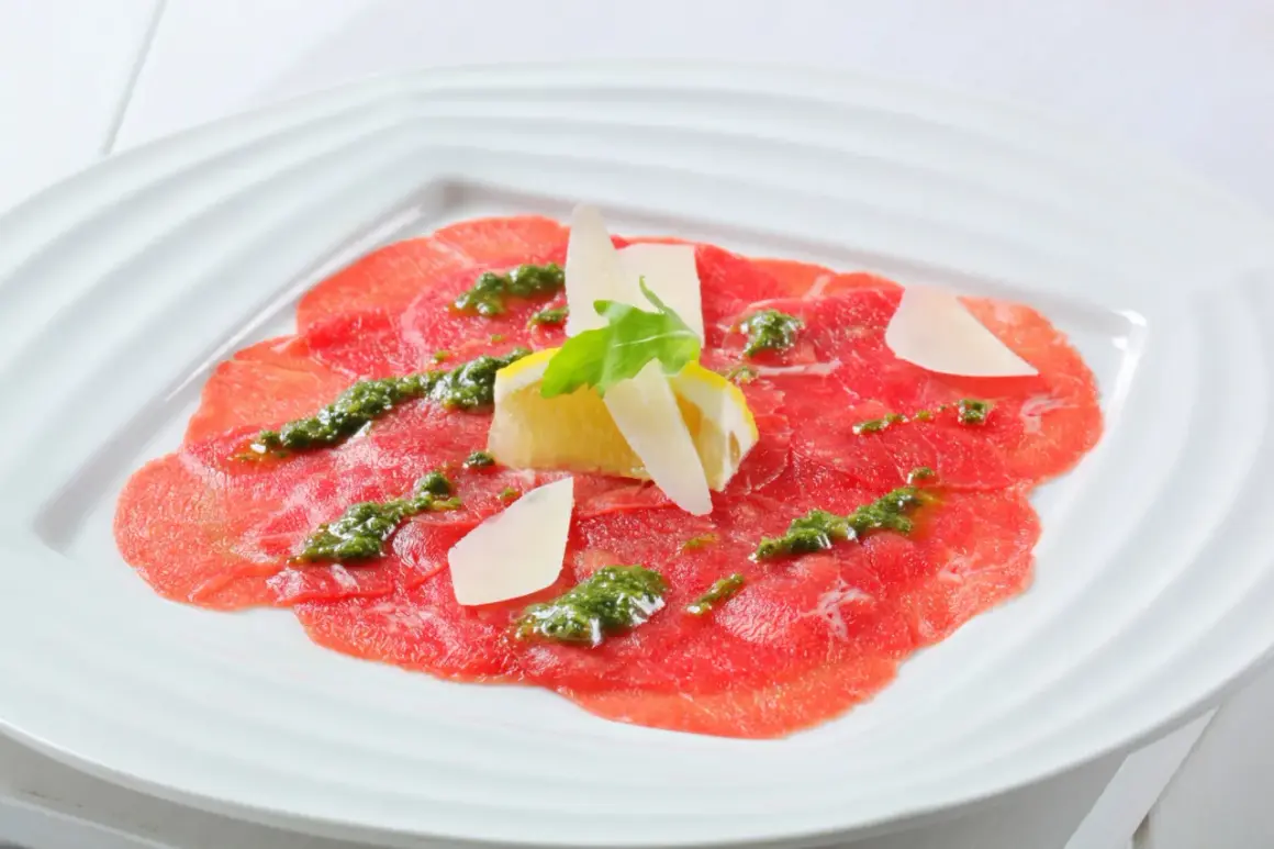 Carpaccio de bœuf finement tranché, arrosé d'une sauce verte et garni de copeaux de parmesan, de citron et d'une feuille de roquette.