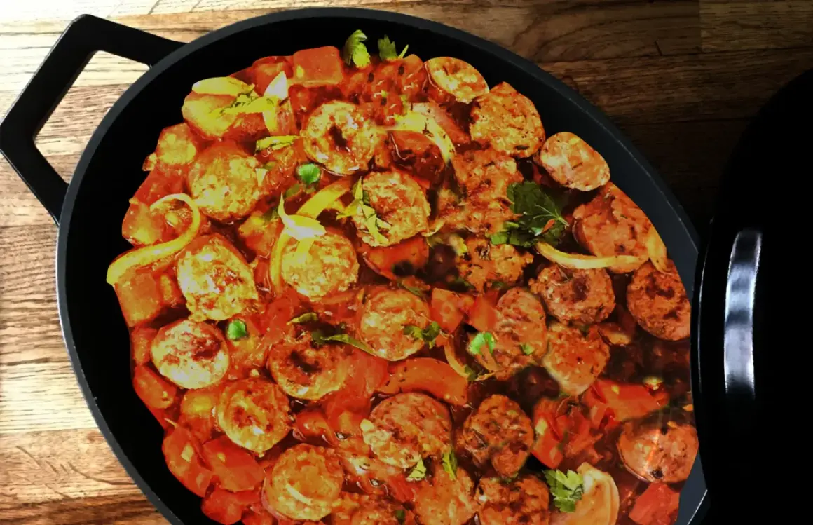 Un plat de rougail saucisse mijote dans une cocotte noire, avec des morceaux de saucisses, des tomates et des oignons.