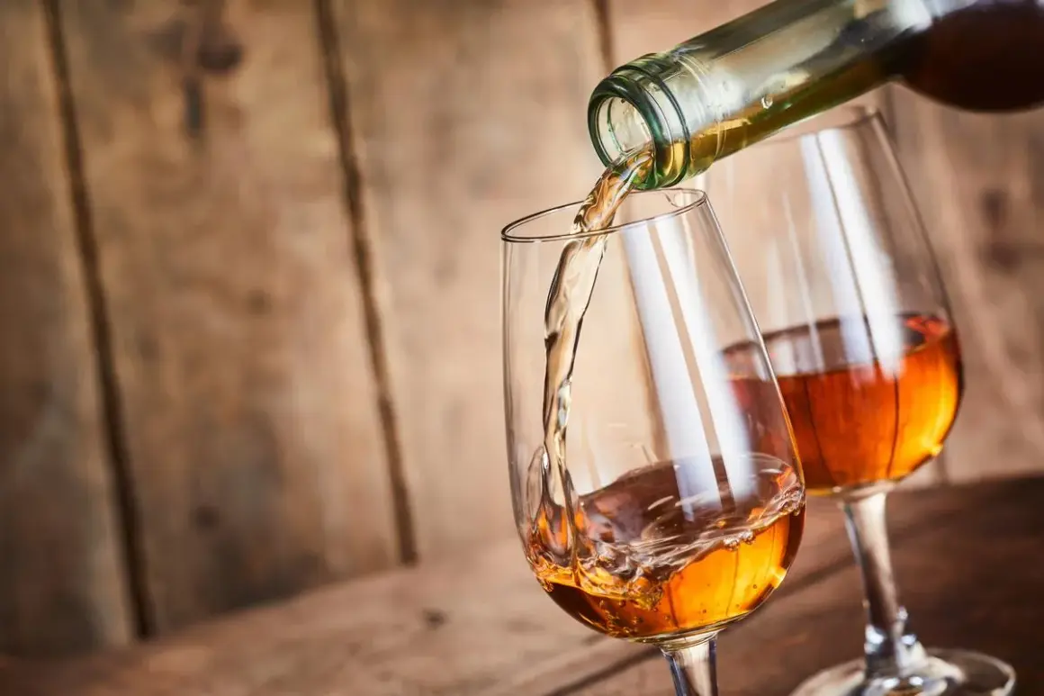 Un verre se remplit d'un vin paillé ambré, un autre verre est déjà servi. Ambiance chaleureuse sur fond de bois.
