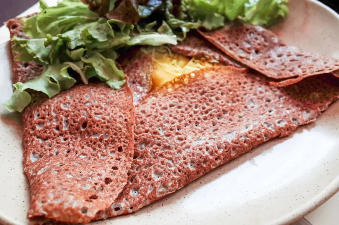Une galette sarrasin pliée, garnie de fromage fondant et de salade verte fraîche, sur une assiette beige.